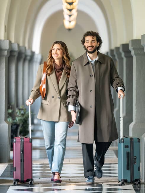 Two guests walk with their luggage down a long arched hallway with a black and white tiled floor.
