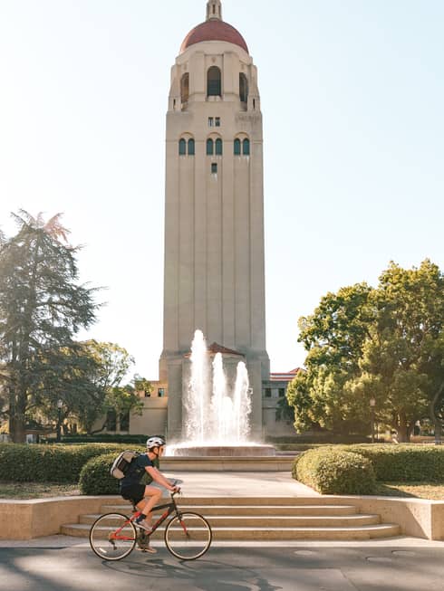 A large tower building with a biker cycling passed it in a park.