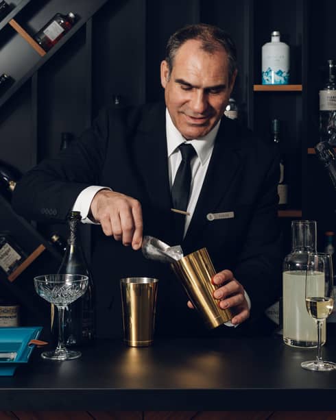 A bartender in suit and tie prepares a drink at a bar counter in an indoor space with dark decor and artistic shelves in the background displaying various bottles of wine and spirits