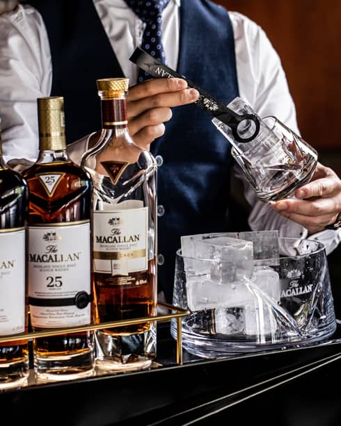 A bartender in tie and vest places a large ice cube in a glass with tongs, next to a five-bottle lineup of Macallan whiskies.