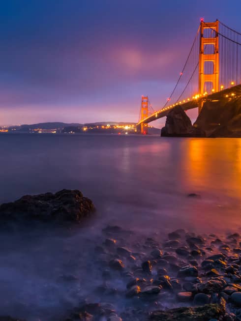 View from the rocky shoreline near the Golden Gate Bridge lit up in vibrant gold and red against a deep blue night sky.