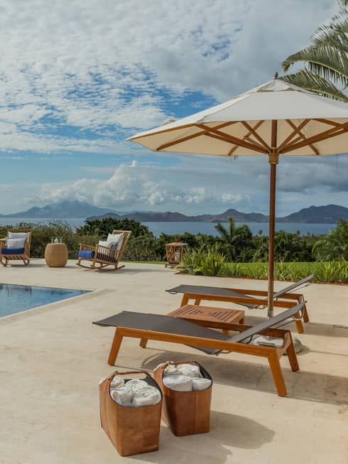 Two lounge chairs under patio umbrella, baskets with towels by outdoor pool