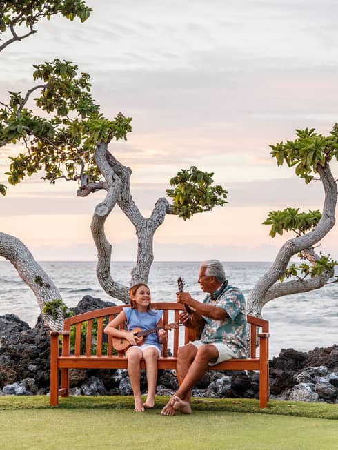 This image depicts a man and a child both playing the ukulele on a bench in front of a tree and the ocean, and is connected to ESG and sustainability