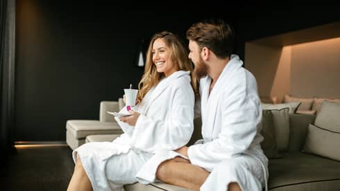 Smiling woman with coffee cup and man in white bathrobes cuddle on large sofa