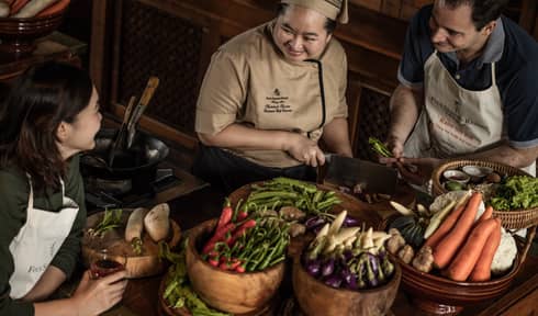  A chef teaching two participants in a cooking class, surrounded by fresh vegetables and ingredients in a rustic kitchen setting.