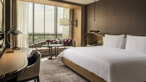 King-sized hotel bed with white linens, modern sofa under sunny window, desk and chair