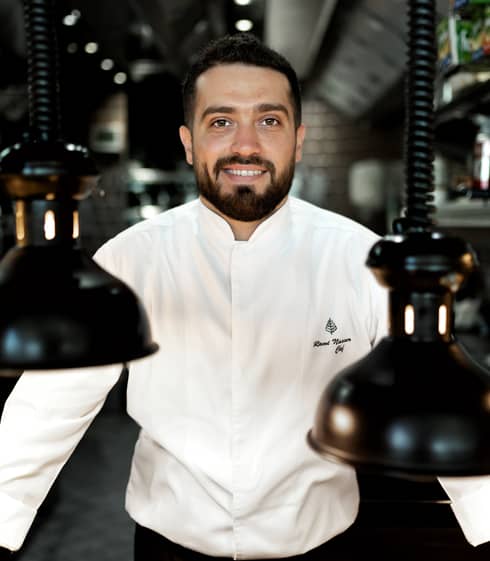 Chef Rami Nasser in his white chef's jacket smiles at the camera, flanked by black vintage pendant lamps hanging from coils.