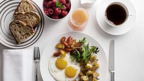 Breakfast plate with sunny-side-up eggs, cherry tomatoes, greens, and roasted potatoes, alongside toast, raspberries, grapefruit juice and coffee.