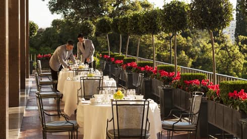 Two servers in grey jackets with black ties set outdoor dining tables with white tablecloths, dishes, red flowers along side