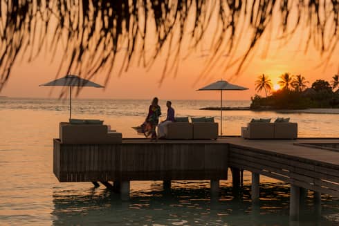 Private island dining at sunset. Woman with long dress blowing in the wind, man sits on the edge of white sofa