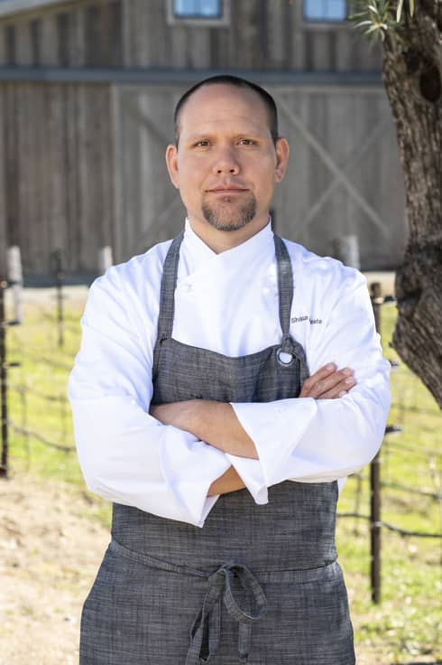 A chef standing outside near a tree and in front of a barn.