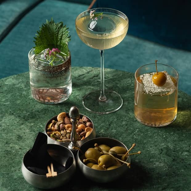 A green marbled table topped with bowls of green olives and nuts and three cocktails in clear glasses