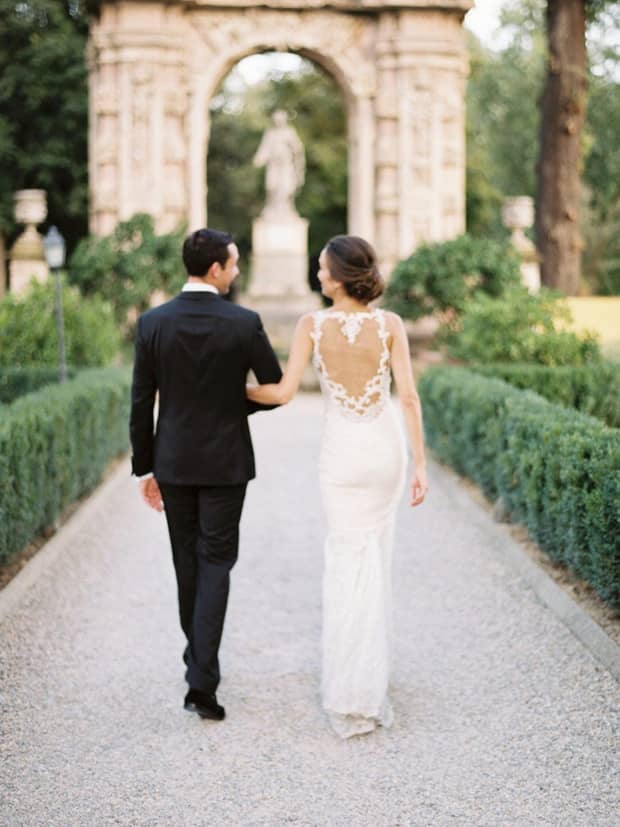 The back of couple walking arm in arm down a pathway, surrounded by greenery, with an elegant stone arch and statue in the background