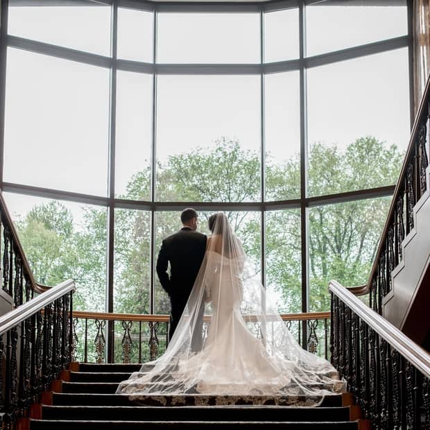 View of a bride and groom from behind standing in a large stairwell facing a wall with floor-to-ceiling windows overlooking a row of trees