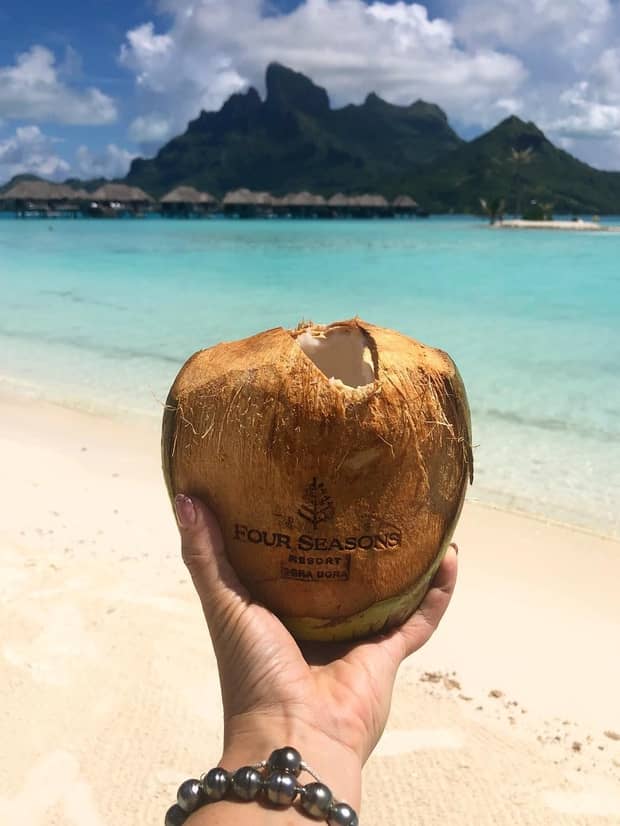 Hand holding coconut with Four Seasons logo and hole for straw against white sand beach, turquoise lagoon