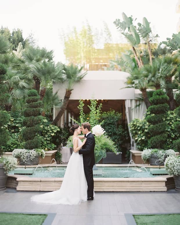 Bride and groom dance on path by fountain, landscaped gardens and palm trees