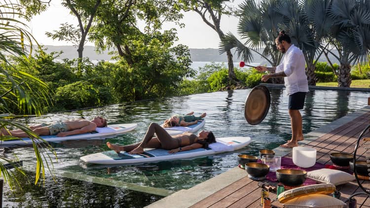 Three people float on stand-up paddleboards in a private pool while someone plays a gong on the deck