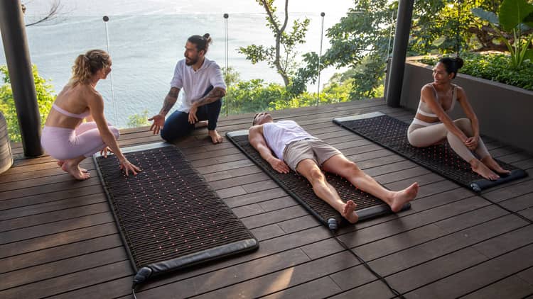 Three people sit and lay on red-light therapy mats as an instructor talks