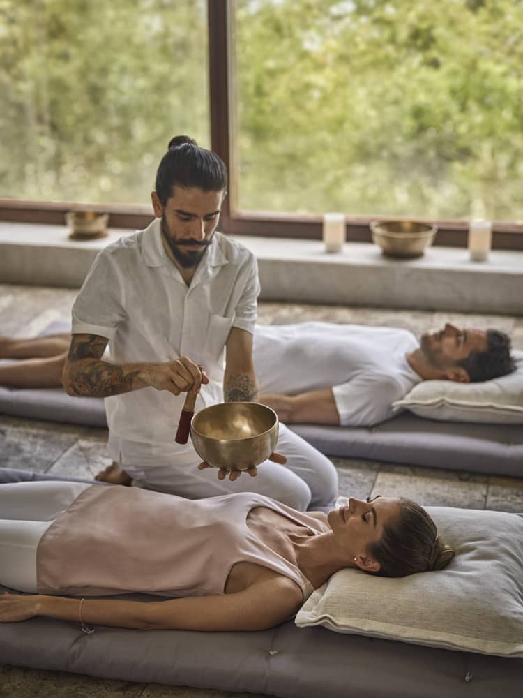 A man in a white short-sleeve button down and white pants is kneeling and playing a sound bowl between a man and a woman who are laying down