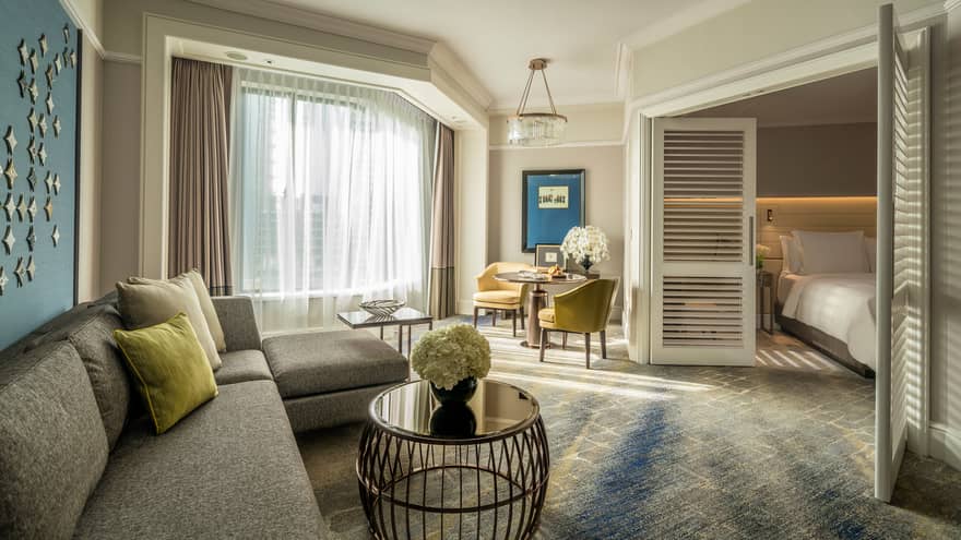 Four Seasons Executive Suite L-shaped sofa by sunny window with white curtains, table, wood shutters