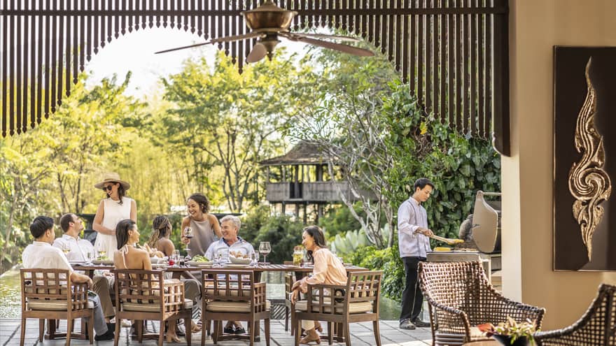 A smiling group around a rustic patio set with wine and appetizers, as a chef barbecues corn and light filters through trees.