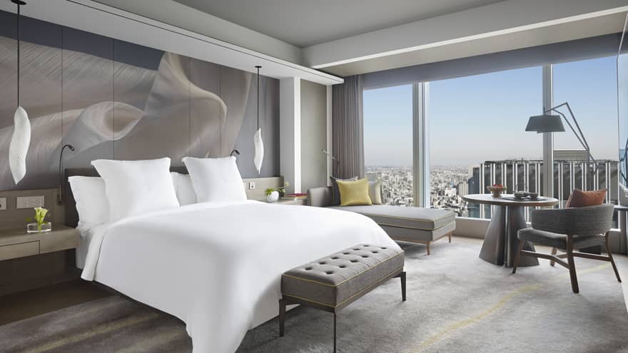 Guest room with king bed, tufted bench, chaise lounge, table with armchair, pendant lights and large window offering a city skyline view