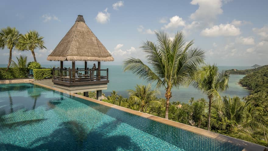 Thatched-roof pavilion at edge of outdoor swimming pool overlooking tropical trees, mountain, ocean