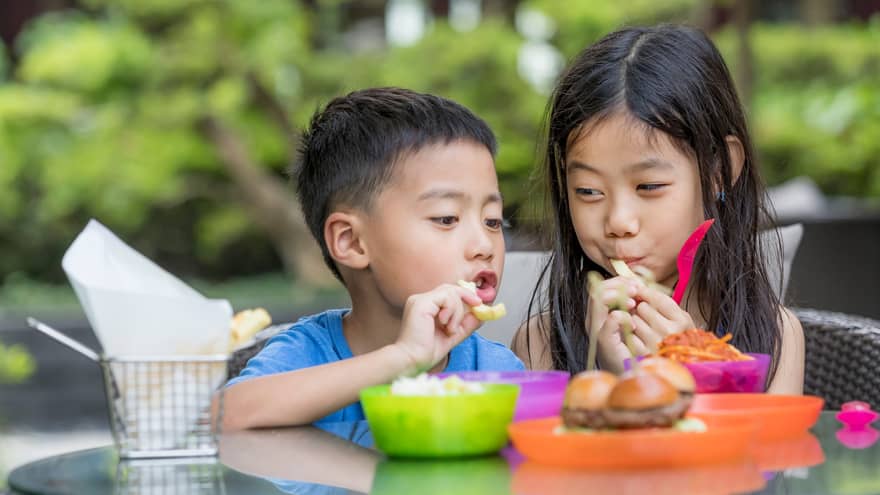 Children enjoying a variety of dishes in colourful plastic dinnerware on a glass-top table on a patio surrounded by greenery.