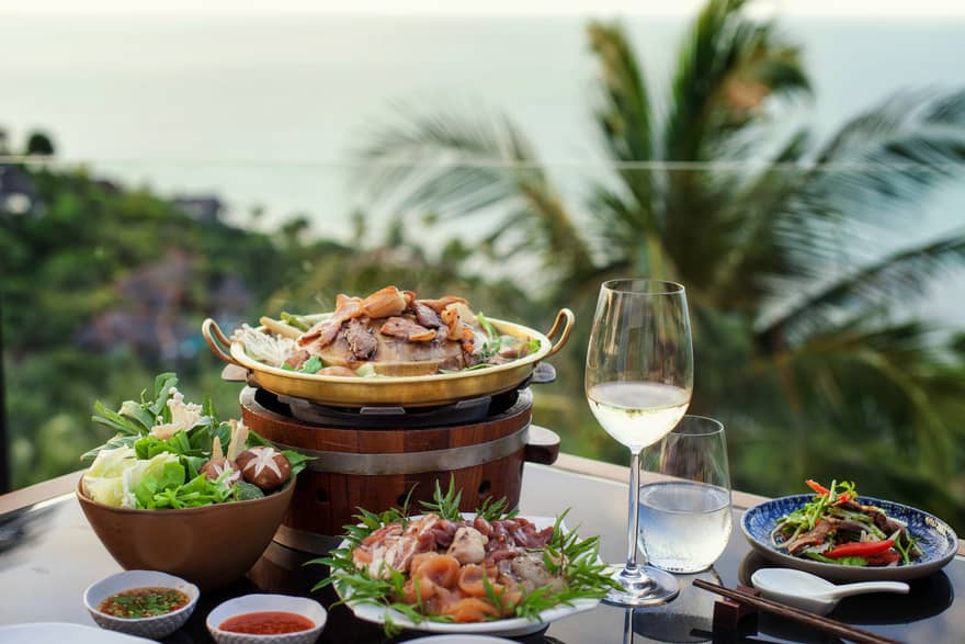 Four plates of food, glass of water and white wine, tropical scenery in background