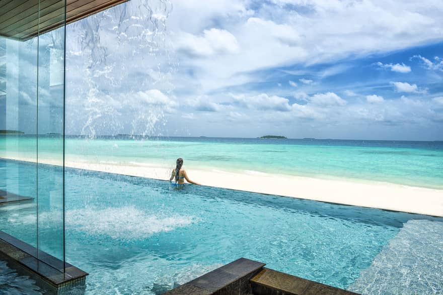 Woman stands at edge of infinity swimming pool looking out over white sand beach, blue ocean