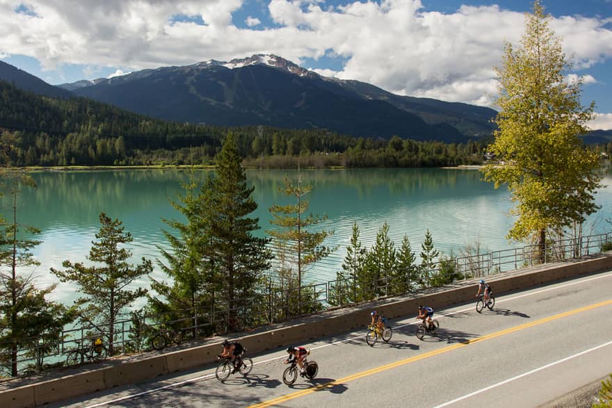 Cyclists bike down road past trees, blue glacier lake under mountains 