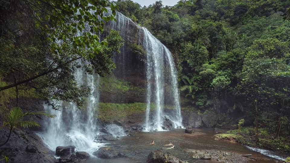 Water cascading over a mossy cliff encompassed by lush greenery; below the waterfall, three people relax in the plunge pool.