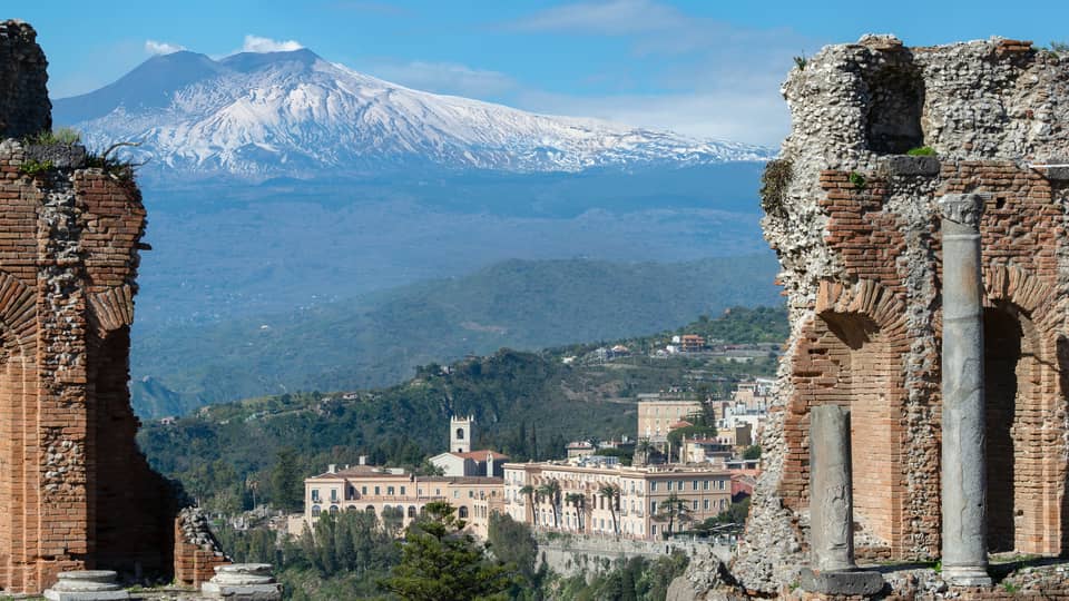View of ancient stone ruins with a distant town and snow-capped mountains in the background.