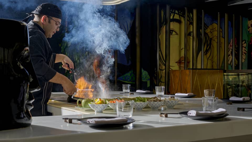 Chef in black coat cooks dish over open flame in show kitchen