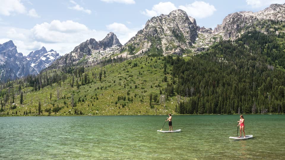 Two people on stand-up paddleboards on lake under mountain hills, trees, snowy peaks