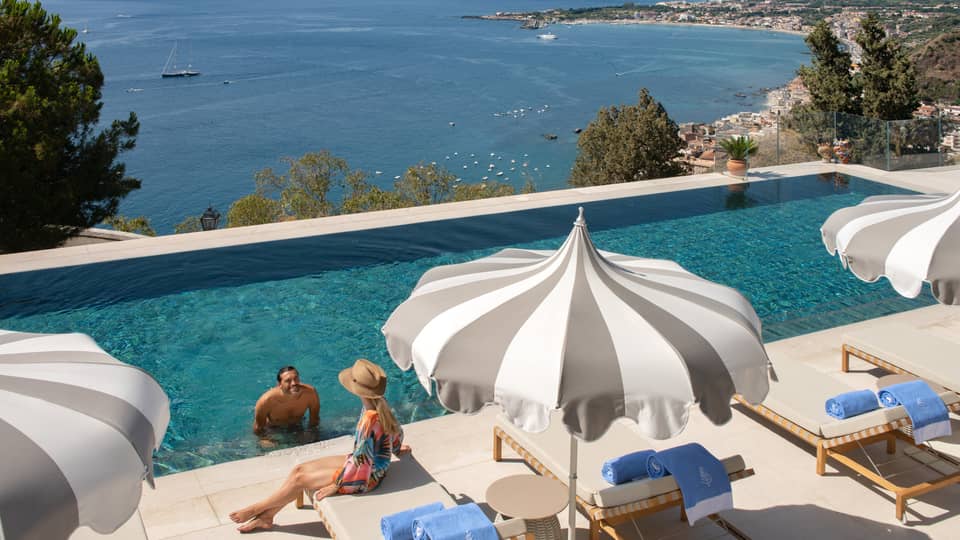 A man at the edge of the infinity pool speaking to a woman sitting on a deck chair