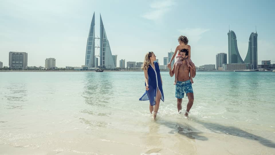 A man with a teenager on his shoulders walks with a woman through shallow ocean water with the city of Bahrain Bay in the background