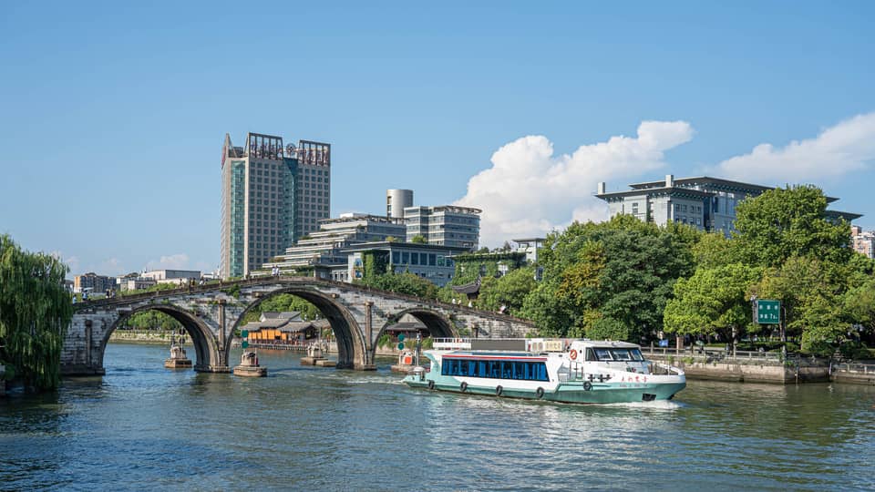 A tour boat passes under the centre arch of a historic stone bridge spanning a canal flanked by trees and modern buildings.