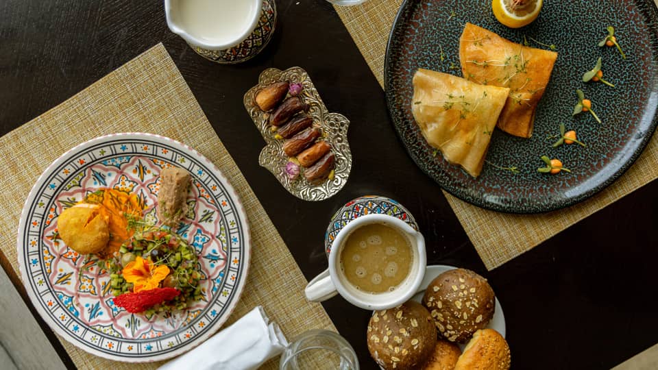 Two plates of international dishes, served with sides of dates and rolls