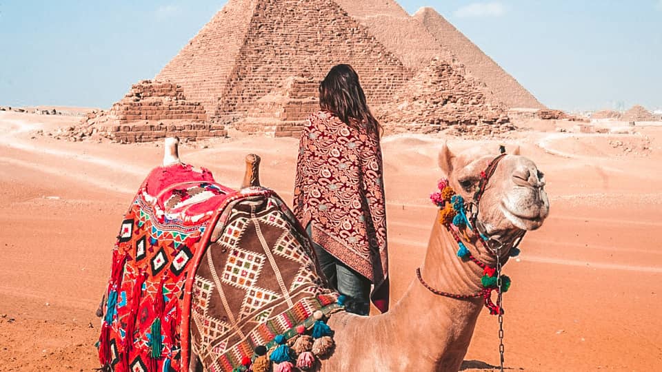 A woman behind a camel covered in colourful blankets looking at pyramids in the distance.