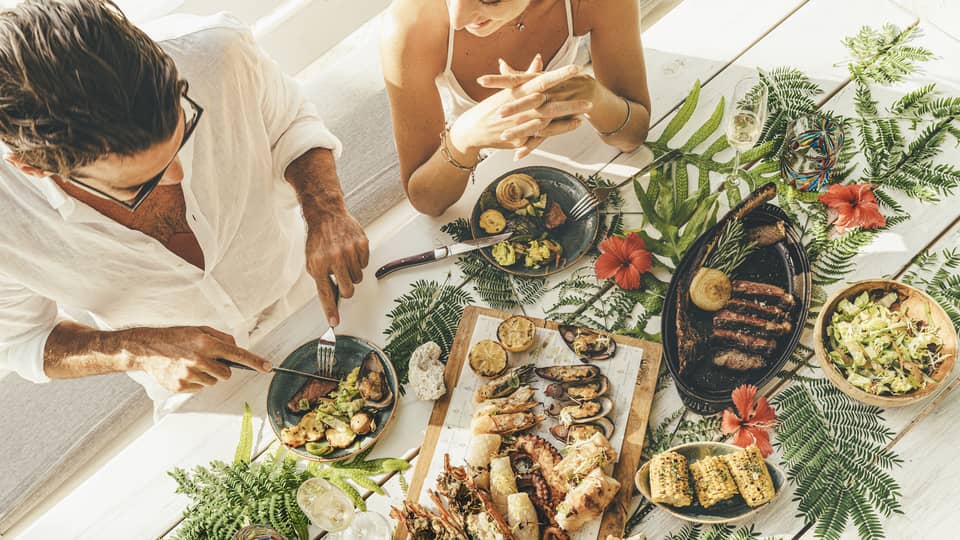 A couple at a table of seafood canapés, grilled meat, corn and champagne flutes, adorned with palm fronds and red hibiscus.