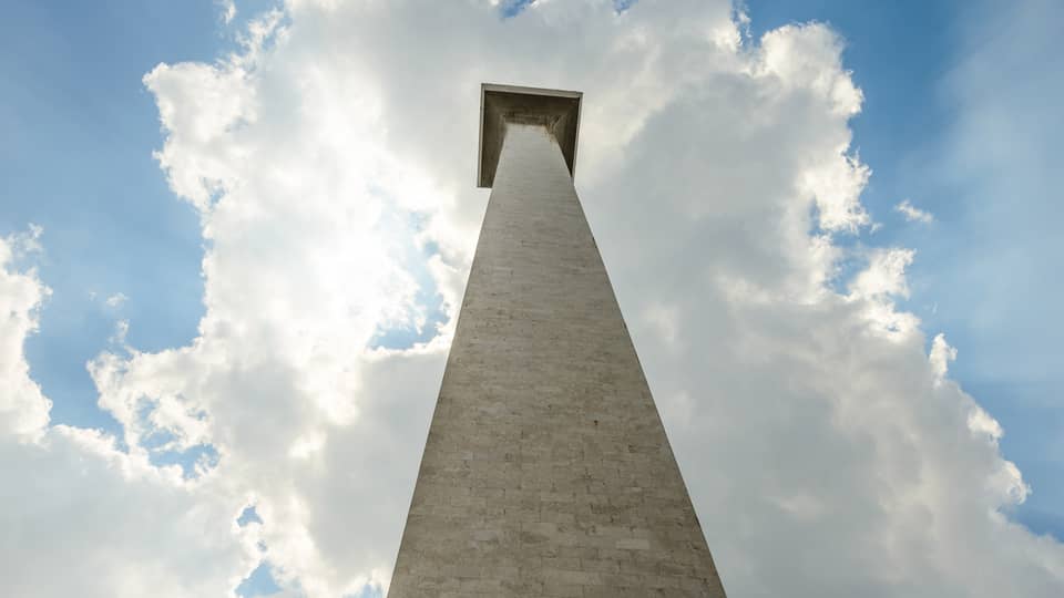 Staring up from the base of the Monas National Monument; a blue sky and the sun peeking through the clouds as a backdrop.