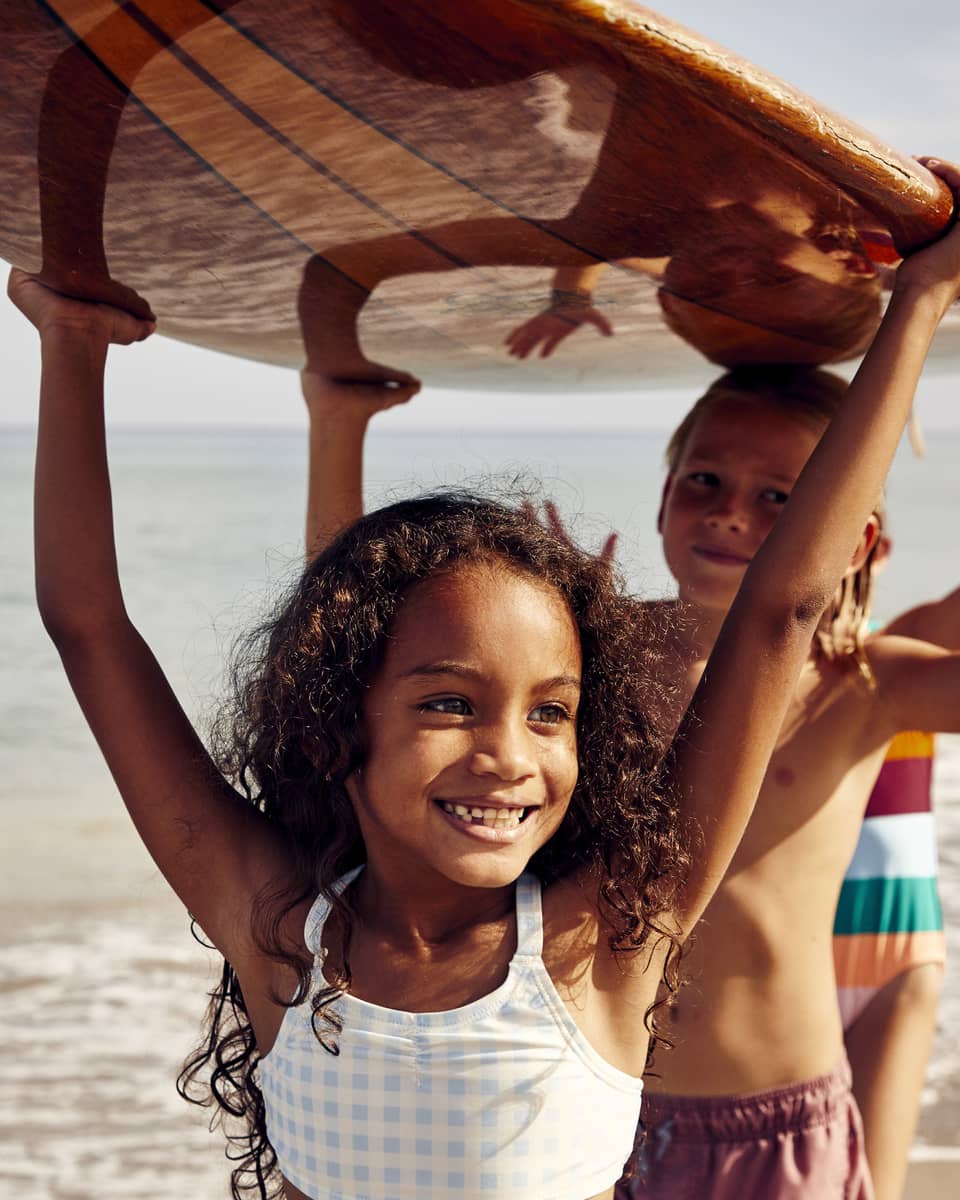 Three young children carry a surfboard on the beach, smiling and enjoying the sunny day. The ocean is in the background.