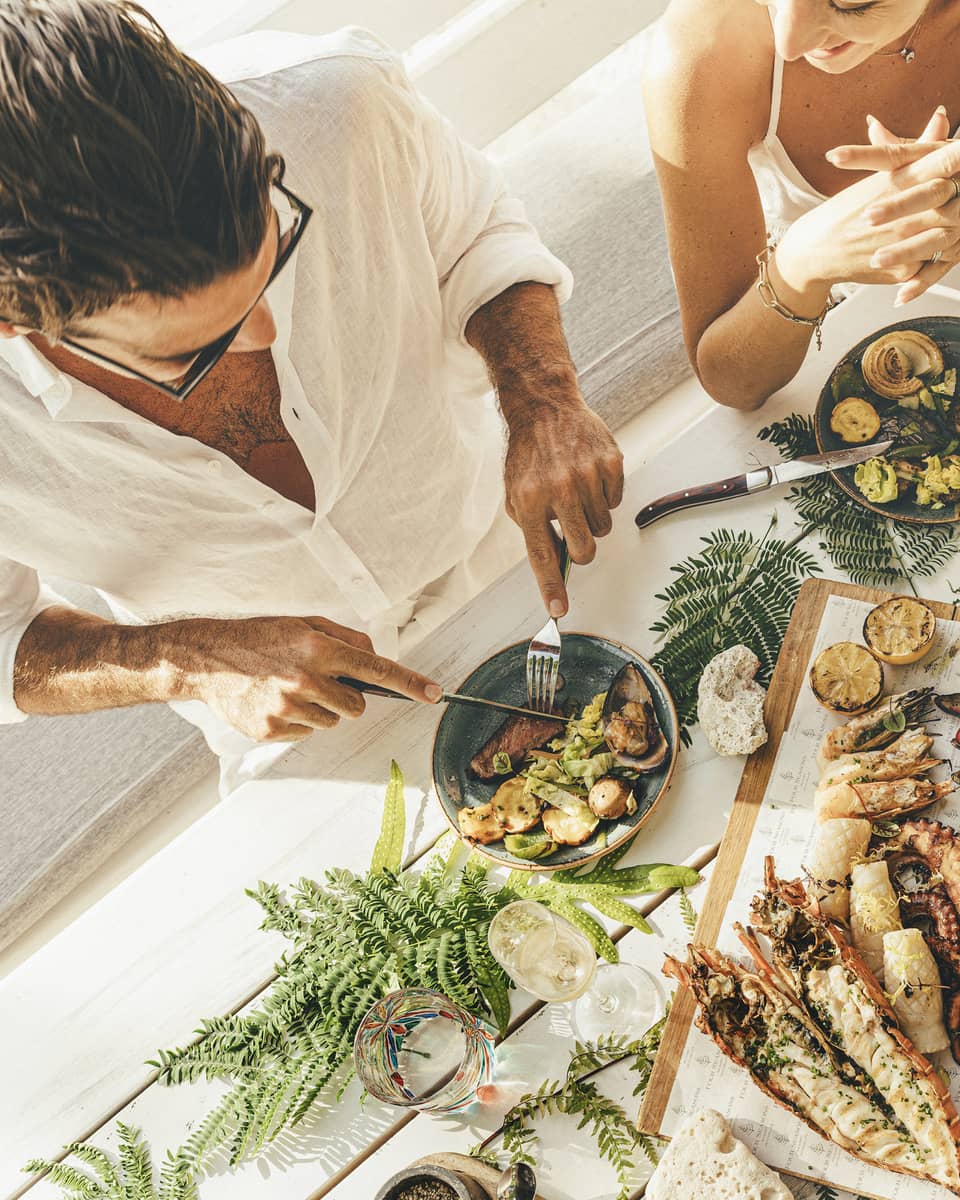 A couple at a table of seafood canapés, grilled meat, corn and champagne flutes, adorned with palm fronds and red hibiscus.