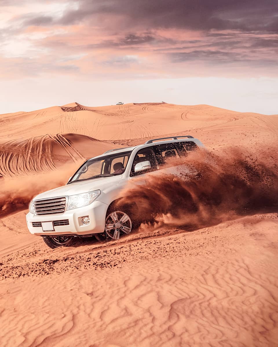 Sand flies from tires of two white SUV's driving across dunes
