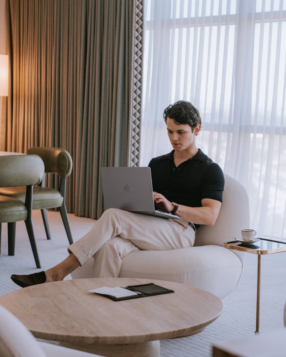 Person sitting in a living room with floor-to-ceiling windows and typing on a laptop.