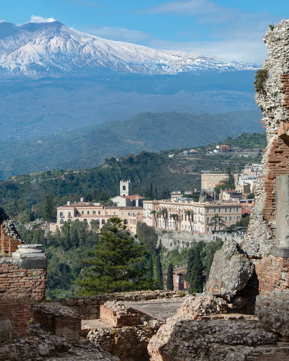 View of ancient stone ruins with a distant town and snow-capped mountains in the background.