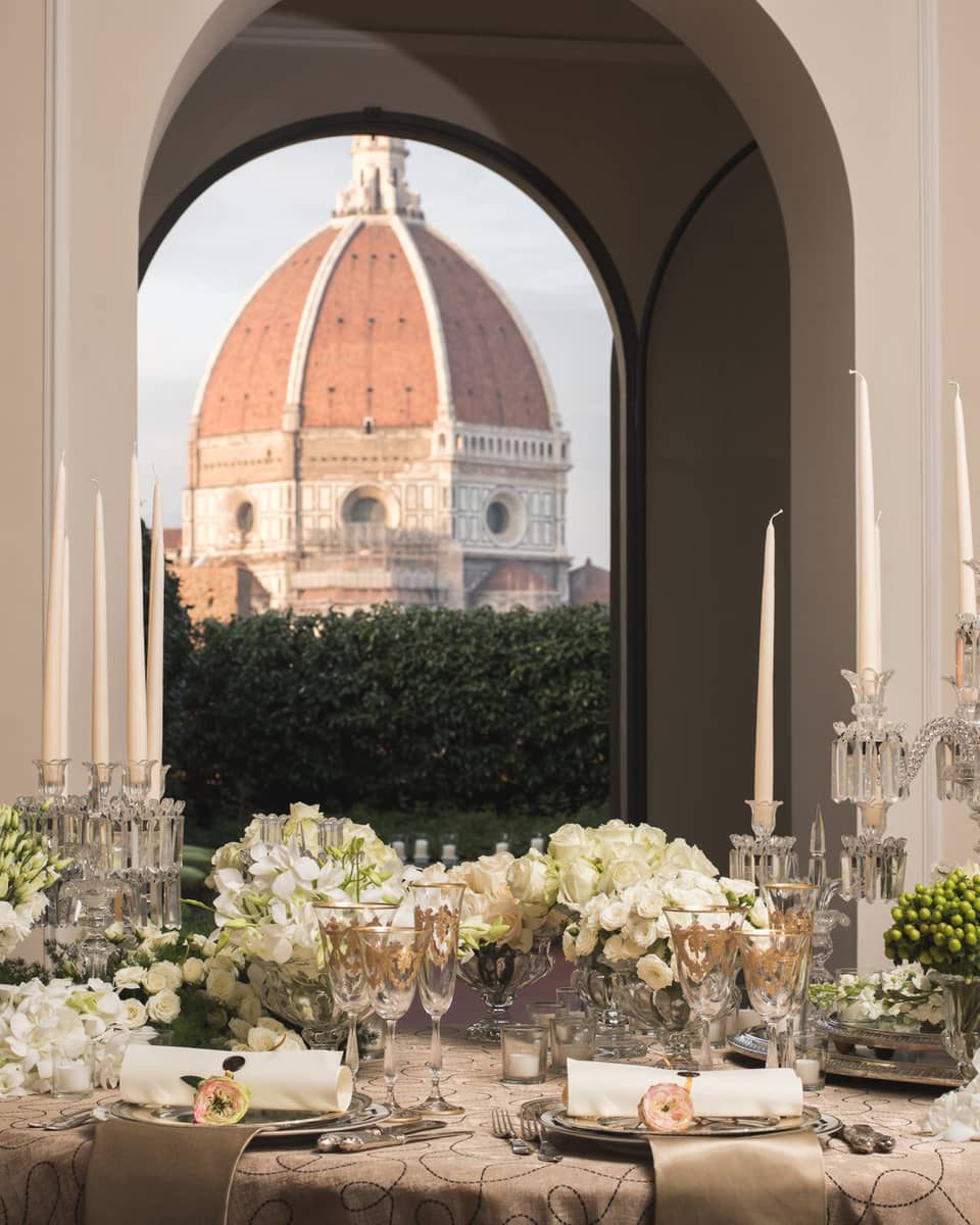 Elegant dining table set with candles, flowers, and glassware, with a large dome visible through arched windows in the background.