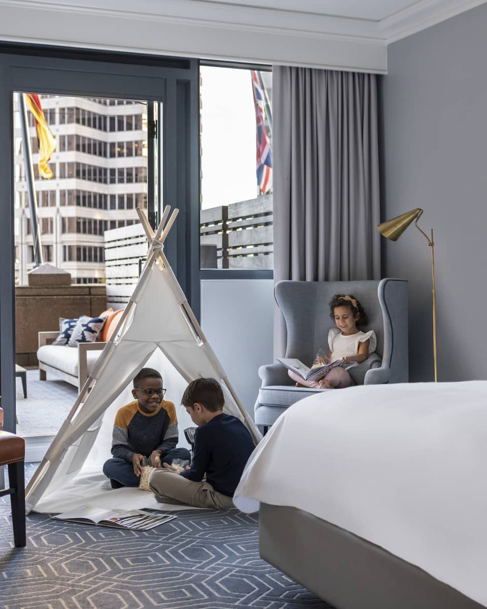 Children sitting in a tent and on a chair in a hotel room.