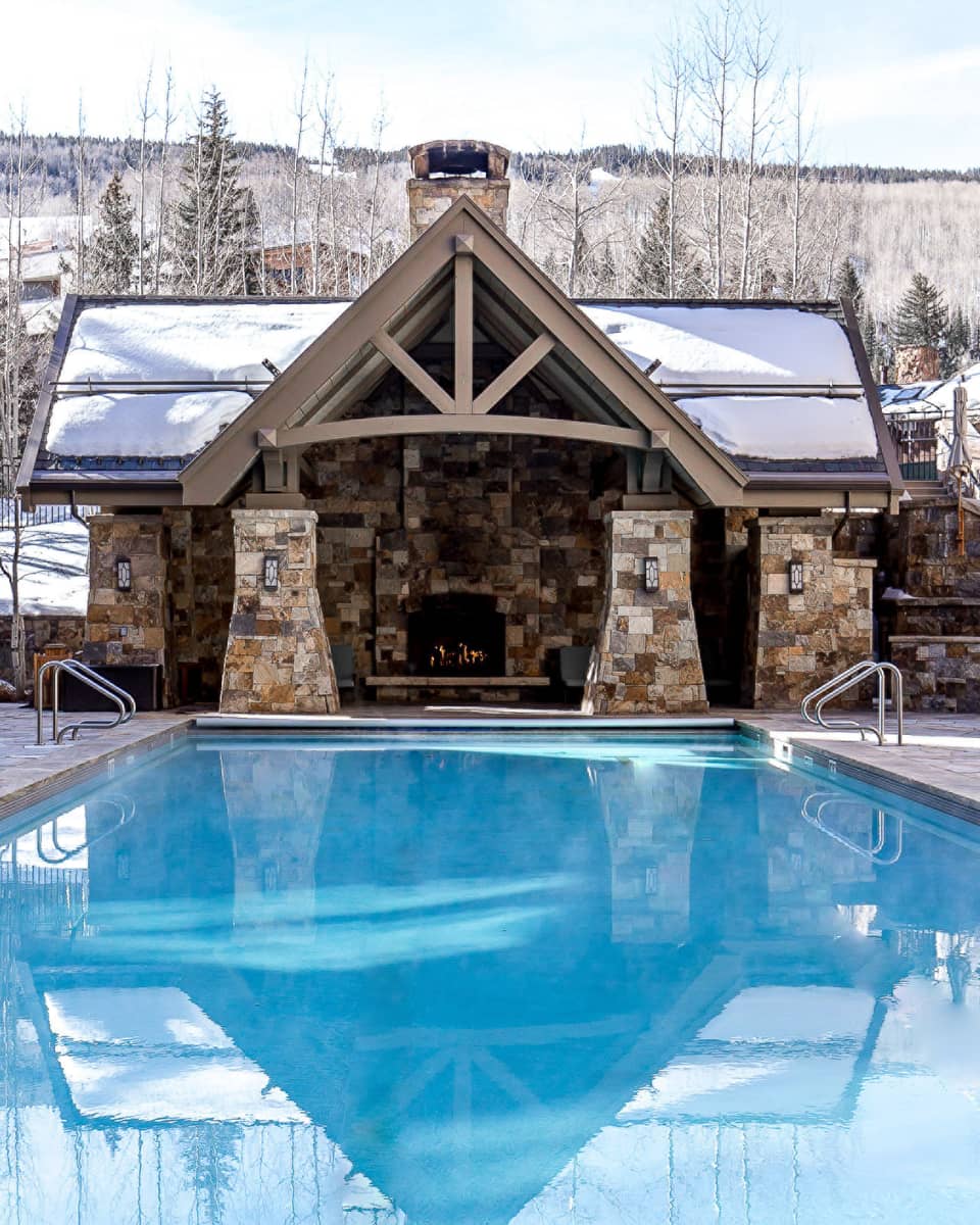 Outdoor pool with lounge chairs, a cabin with a fireplace and pine trees in the background.